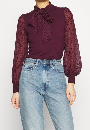 Vrouw draagt een paarse blouse met doorzichtige gebobbelde mouwen met stippen en een strikjeskraag, gecombineerd met een lichtblauwe high-waisted jeans.