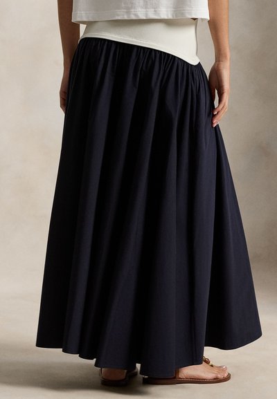 Μακρύ navy βαμβακερό φόρεμα με συγκεντρωμένη μέση, με ρέον σχέδιο. Φορώντας το με ταμπά σανδάλια σε ουδέτερο υφασμάτινο φόντο.