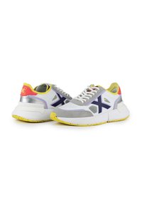 Baskets de sport blanches avec des accents en daim gris et en maille, logo violet et semelle tachetée de jaune. Présente des détails métalliques argentés et roses.