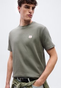 Νέος άνδρας που φοράει ελιά-πράσινη υφασμάτινη μπλούζα με μικρό λογότυπο, με το χέρι στην τσέπη και καρό πουκάμισο δεμένο γύρω από τη μέση, κοιτάζοντας πλάγια.