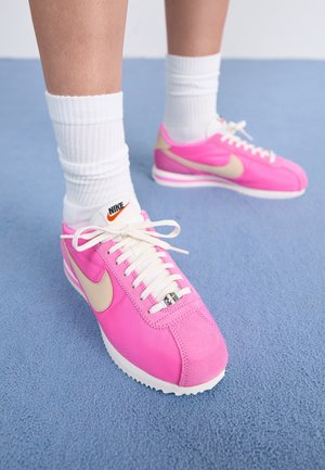 Personne portant des baskets Nike rose vif avec un swoosh beige et des lacets blancs, assorties à des chaussettes blanches côtelées à la cheville sur un sol bleu.