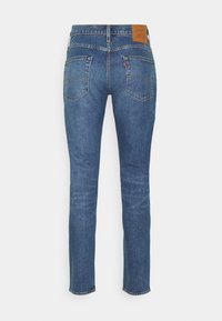 Jean en denim bleu à jambes droites vu de dos avec deux poches plaquées et une étiquette de marque en cuir sur la ceinture.