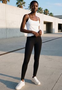 Woman wearing a white tank top, black leggings, and white sneakers standing outdoors on a concrete surface with palm trees in the background.