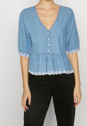 Nainen pukeutuneena vaaleansiniseen nappiliseen puseroon, jossa on liehukkeet hihansuissa ja valkoista kukkaompeletta, yhdistettynä mustiin housuihin.