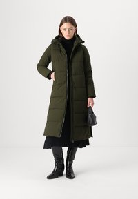 Lang, groen pufferjack met een capuchon, voorzien van een ritssluiting en een gestikte patroon. Gecombineerd met een zwarte coltrui en kniehoge zwarte laarzen.
