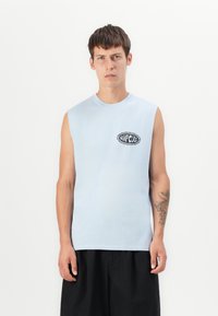 Ung man med kort hår bär en ljusblå ärmlös tröja med "Rip Curl"-logotyp och svarta shorts, stående framför en vit bakgrund.