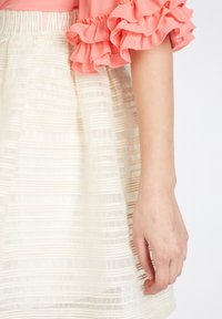 Brazo de una persona que lleva una blusa de manga rizada rosa y una falda blanca texturizada con patrones de rayas horizontales.