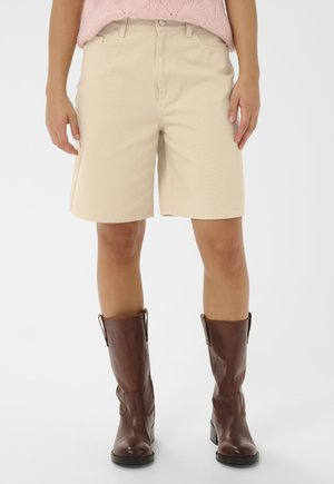 Person iført beige knælange shorts, brune læderstøvler til midt på læggen og en lyserød strikket sweater, stående mod en hvid baggrund.