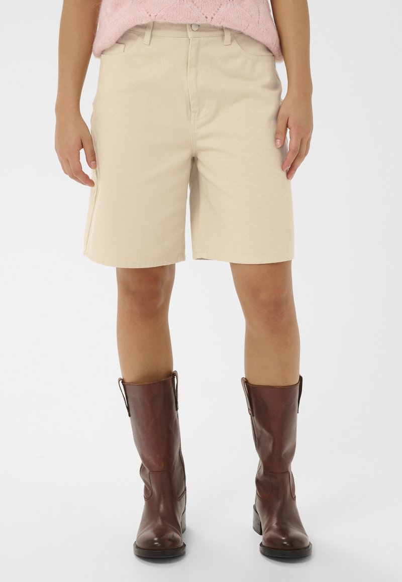 Personne portant un short beige jusqu'aux genoux, des bottes en cuir marron mi-mollet et un pull en tricot rose, debout devant un fond blanc.