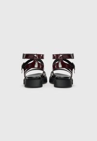 Sandales en cuir verni bordeaux avec un dos ouvert, une sangle à boucle et une semelle en caoutchouc noir. Présente des lanières délicates et un design épuré.
