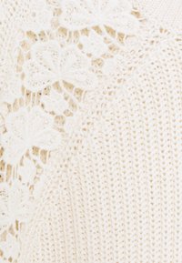 Sweter dziany w kolorze kremowym z kwiatowymi koronkowymi detalami, charakteryzujący się fakturowanym wzorem i miękką przędzą. Gładka powierzchnia z misternymi akcentami szwów.