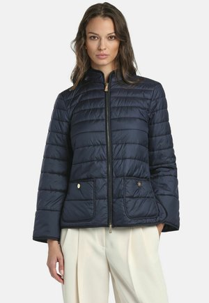 Nő, aki sötétkék, steppelt cipzáros dzsekit visel két első zsebbel, magas derekú fehér pliszírozott nadrággal, fehér háttér előtt áll.