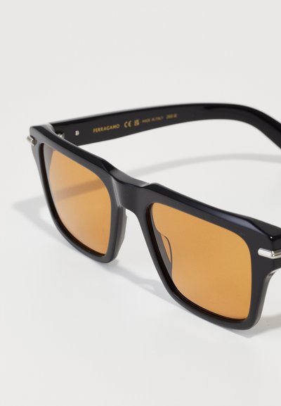 Lunettes de soleil noires surdimensionnées avec des verres bruns. Cadre en plastique solide avec des accents en métal sur les charnières. Conception angulaire.