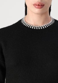 Crna pletena veste s teksturiranim završetkom, s kontrastnim bijelim i crnim prugastim rebrastim izrezom. Prikaz izbliza područja ovratnika.