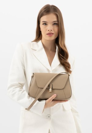 Vrouw met lang bruin haar in een wit colbert houdt een beige leren handtas met gouden sluiting en schouderriem vast tegen een witte achtergrond.
