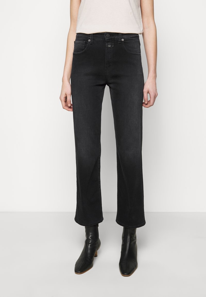 Personne portant un jean noir droit et des bottines noires, debout devant un fond clair uni.