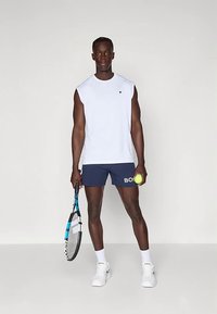 Λευκό αθλητικό τοπ χωρίς μανίκια, σκούρο μπλε σορτς με το λογότυπο "BOSS", κρατώντας ρακέτα τένις και μπάλα, φοράει λευκά αθλητικά παπούτσια και κάλτσες.