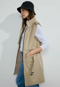 Beige ärmellose Weste mit hohem Kragen, Reißverschluss vorne und seitlichen Taschen. Besitzt eine taillierte Taille und graue Seitenpaneele.