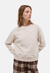 Sweatshirt rose clair avec un col rond et des manches longues, présentant une texture mouchetée. Associé à un pantalon à carreaux marron.