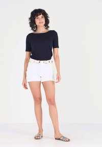 Mørkeblå kortermet topp, hvite shorts med belte og mønstrede sandaler. Modellen står mot en ensfarget bakgrunn og viser frem antrekket.