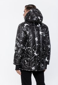 Mężczyzna ubrany w czarną kurtkę puchową z białym wzorem plam farby, z kapturem na głowie, widoczny od tyłu na jednolitym białym tle.