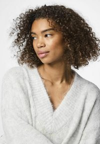 Balta, pūkuota megzta palaidinė su giliu V formos iškirpimu ir raukšlėtu dekoru. Modelis turi garbanotus plaukus ir yra neutralioje aplinkoje.