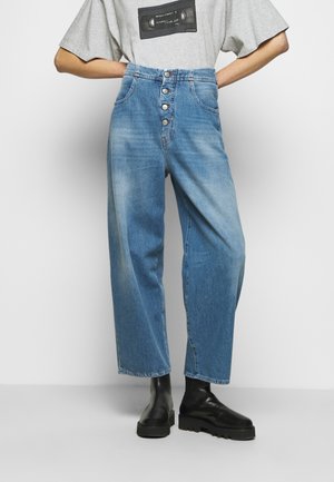 Persona con pantalones vaqueros azules de talle alto y pierna ancha con cierre de botones, camiseta gris con estampado de casete, y botas negras de plataforma gruesa.