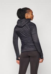 Giacca nera isolante con cappuccio, caratterizzata da un design trapuntato e maniche lisce, abbinata a leggings neri. Superficie testurizzata e forma aderente.