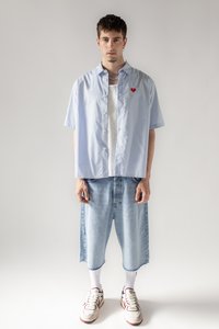 Lichtblauw gestreept shirt met korte mouwen en een rode hartdetail, gedragen over een witte tanktop, gecombineerd met lichtblauwe baggy spijkerbroekshorts en sneakers.