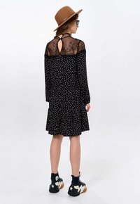 Zwart jurkje met stippen, lange mouwen, kanten details op de rug, gerimpelde zoom, en gecombineerd met zwarte hoge sneakers en een bruine hoed.