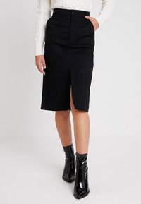 Musta polvipituinen hame, jossa edessä halkio, muotoiltu vyötärönauha ja syvät taskut, yhdistetty kiiltäviin mustiin nilkkureihin. Sileä kangaspinta.