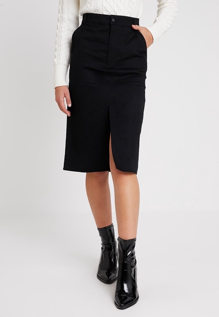 Musta polvipituinen hame, jossa edessä halkio, muotoiltu vyötärönauha ja syvät taskut, yhdistetty kiiltäviin mustiin nilkkureihin. Sileä kangaspinta.