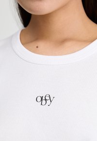 T-shirt bianca a coste con collo rotondo, caratterizzata da un piccolo logo nero che recita "obey" centrato sulla zona del petto.