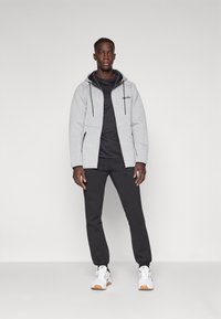 Graue Zip-Hoodie mit schwarzen Akzenten, über einem schwarzen Shirt getragen, kombiniert mit schwarzen Hosen und weißen Sneakern. Glatte Textur, lässige Passform.
