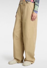 Pantalones de pierna ancha en color beige, confeccionados en un algodón resistente. Cuentan con dos bolsillos frontales, una cintura alta con detalle de botón y una tela texturizada.