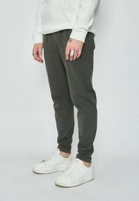 Personne portant un pantalon de jogging gris foncé, un sweatshirt blanc, des baskets blanches et des bagues aux doigts, se tenant sur un fond clair.
