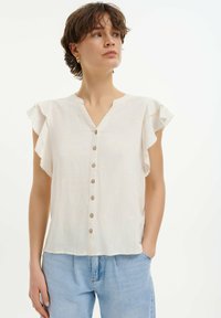 Blusa corta beige chiaro con maniche corte e scollatura a V, dotata di ricami floreali decorativi e chiusura con bottoni, realizzata in tessuto leggero.