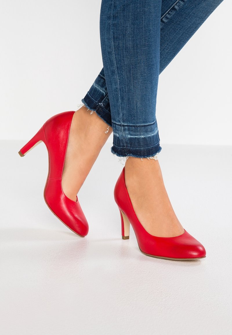 Pieds chaussés de chaussures rouges à talons hauts associés à un jean skinny bleu à ourlet effiloché, sur fond blanc uni.