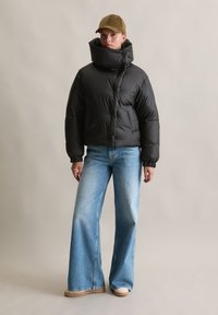 Melnā pūšu jaka ar augstu apkakli, priekšējiem pogām un sānu kabatām, apvienota ar gaiši zilām platām biksēm un bēšām kurpēm.