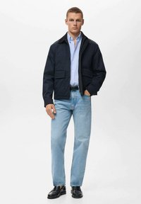 Chaqueta navy con botones en la parte frontal y dos bolsillos en el pecho, llevada sobre una camisa azul clara, combinada con jeans azul claro y zapatos negros.