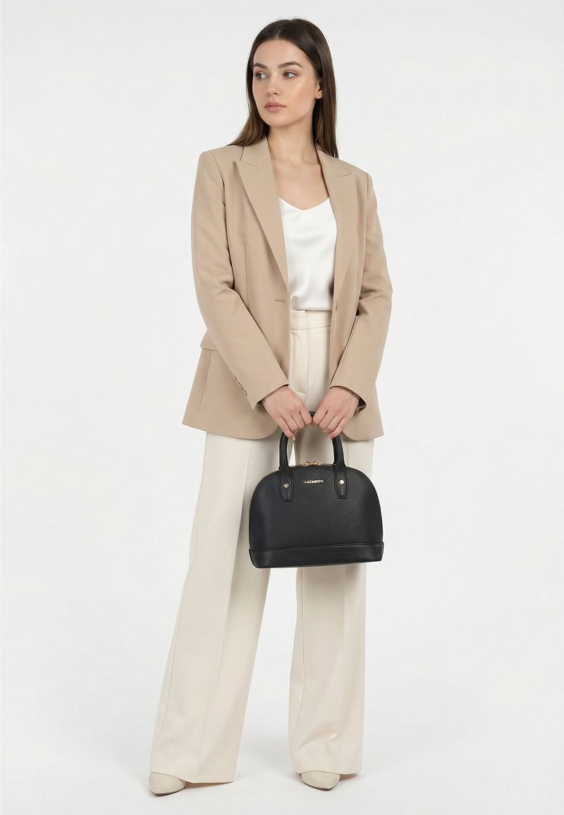 Mujer con blazer beige y pantalones de pierna ancha sosteniendo un bolso negro, de pie frente a un fondo blanco liso, mirando hacia un lado.