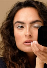 Vrouw met golvend donker haar brengt make-up aan met een kleine zwarte spiegel. Ze heeft een licht zilveren oogschaduw en een natuurlijke lipkleur, met gedefinieerde wenkbrauwen.