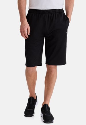 Mann trägt schwarze Shorts mit elastischem Bund und schwarze Sportschuhe, steht vor weißem Hintergrund.