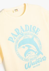 Cremefarvet bomulds T-shirt med en struktureret overflade. Har et blåt grafisk motiv af en delfin og ordene "PARADISE Waves" i fed skrift.