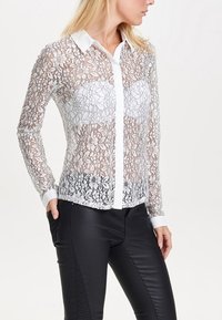 Kanten shirt met lange mouwen, ingewikkeld bloemenpatroon, witte kraag en een sluiting met knopen aan de voorkant, gecombineerd met zwarte leren broek.