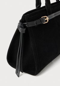 Crna suede torba sa strukturiranim oblikom, koja ima sjajnu crnu kožnu ručku i zlatni detalj kopče sa strane.