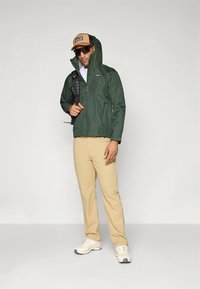 Zelena vodoodporna jakna z visokim ovratnikom, bež hlače in beli športni copati. Črn nahrbtnik in rjava kapa z logotipom. Nošene sončna očala.