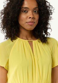 Gelbe Bluse mit gerafftem Ausschnitt und kurzen, fließenden Ärmeln. Weicher Stoff sorgt für einen sanften Fall mit einem dezenten Schlüsselloch-Detail an der Vorderseite.