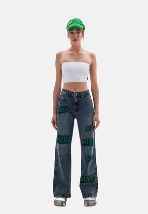 Femme portant une casquette verte texturée, un top tube blanc, un jean large foncé avec des patchs de texte verts et des baskets blanches sur un fond uni.