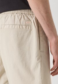 Shorts bej cu talie elastică, confecționați dintr-un material ușor, având un buzunar lateral, un buzunar pe spate cu etichetă și o textură netedă.